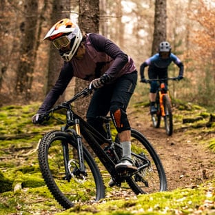 Two mountain bikers riding through a forest trail with moss-covered ground