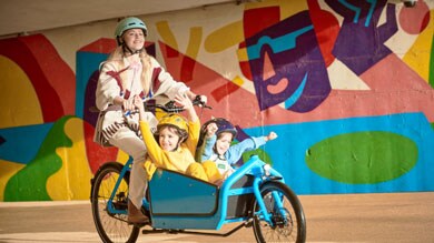 Person fährt ein Lastenrad mit zwei Kindern, bunte Wandmalerei im Hintergrund.