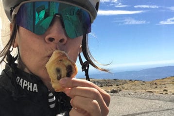 Radfahrer mit Helm und Sonnenbrille isst Gebäck auf einem malerischen Bergpfad.
