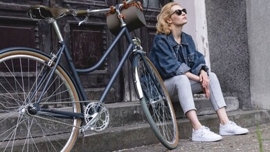 Osoba siedząca obok zabytkowego roweru, w okularach przeciwsłonecznych i kurtce jeansowej.