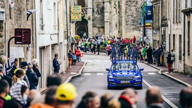 Voiture d’assistance bleue avec vélos dans une rue de village bondée.