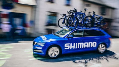 Voiture Shimano bleue avec vélos sur le toit passant devant des bâtiments.