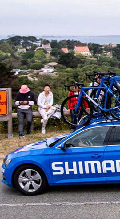 Voiture Shimano bleue avec vélos passant devant des spectateurs au bord de la mer.