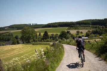 Fietser op een landelijk onverhard pad met velden en bomen onder een heldere hemel.