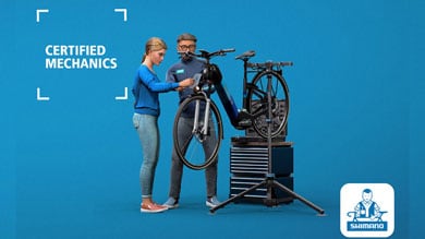 Two mechanics working on a bicycle in a repair shop with text Certified Mechanics and Shimano logo.