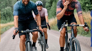 Trzech rowerzystów jedzie szutrową drogą przez zalesiony teren.   1/2