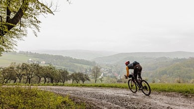 Rowerzysta na szutrowej ścieżce w pagórkowatym terenie z drzewami i pochmurnym niebem.