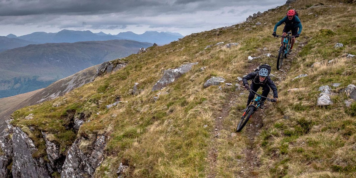 Two mountain bikers descending a narrow trail on a rugged hillside with a scenic valley backdrop