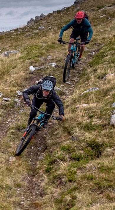 Two mountain bikers descending a narrow trail on a rugged hillside with a scenic valley backdrop