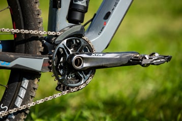 Close-up of a bicycle chainring, crank, and pedals against a grassy background