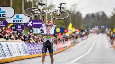 Cyclist lifting bike overhead at finish line, cheering crowd in background.