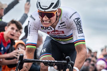 Cyclist in racing attire pushing vigorously while crowd cheers at an outdoor event.