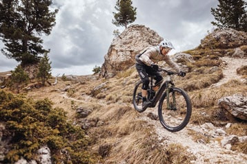Radfahrer mit Helm und Schutzkleidung fährt mit einem Mountainbike auf einem steinigen Abhang in alpiner Landschaft bei bewölktem Himmel.