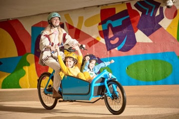 Person fährt ein Lastenrad mit zwei Kindern vorne, bunte Wandmalerei im Hintergrund.