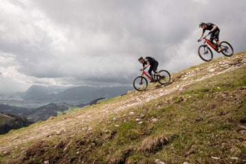Zwei Radfahrer fahren eine grasbewachsene Bergseite unter bewölktem Himmel hinunter.