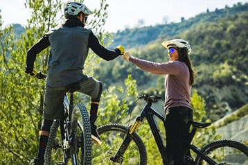 Zwei Radfahrer geben sich auf einem Weg mit Bäumen und Bergen im Hintergrund die Faust.