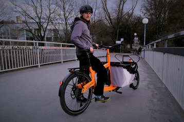 Person auf einem orangefarbenen Lastenrad mit Helm auf einer Brücke.