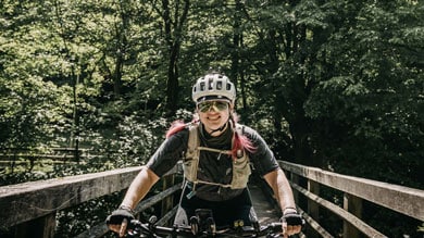 Radfahrer auf einer Holzbrücke im Wald, mit Helm und Sonnenbrille.
