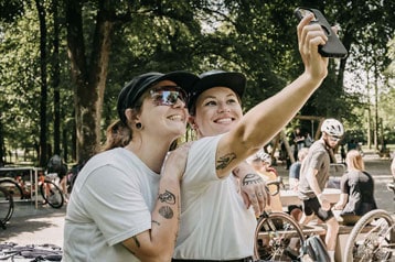 Zwei Personen machen ein Selfie bei einer Outdoor-Veranstaltung mit Fahrrädern im Hintergrund.