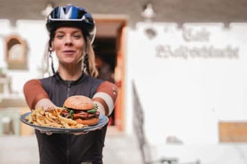 Person bietet einen Burger mit Pommes auf einem Teller an, trägt Fahrradhelm.