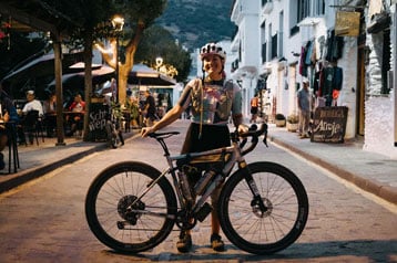 Radfahrer posiert mit Fahrrad auf einer Dorfstraße mit Cafés und Geschäften.