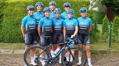 Acht weibliche Radsportlerinnen in blauen und schwarzen Shimano-Teamtrikots posieren für ein Gruppenfoto mit einem blauen Rennrad vor einer grünen Hecke.