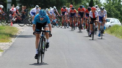 Eine Radfahrerin im blauen Trikot führt das Rennen an und setzt sich von der Verfolgergruppe ab. Das Peloton folgt dahinter mit Fahrern in roten, weißen und blauen Trikots.