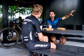 Two cyclists in casual conversation at an outdoor table with bikes nearby.