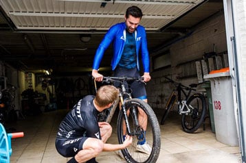 Two cyclists adjusting a mountain bike in a garage.