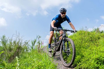 Cyclist riding a mountain bike on a grassy trail under a blue sky.