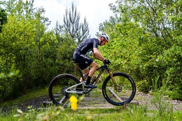 Cyclist in black gear riding a mountain bike on a trail surrounded by trees.