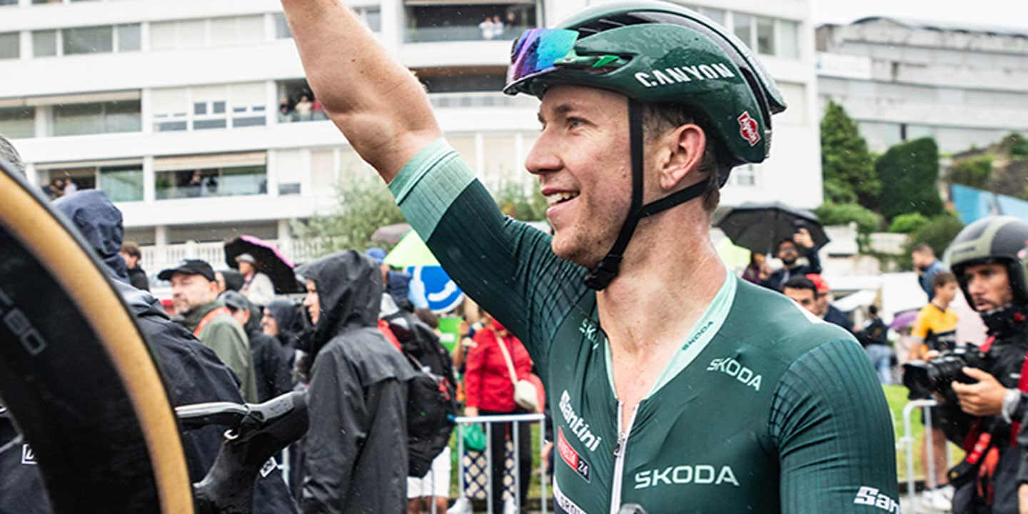 Cyclist in green jersey celebrating with raised arm amid a crowd, holding a bicycle wheel, wearing a helmet and sunglasses.