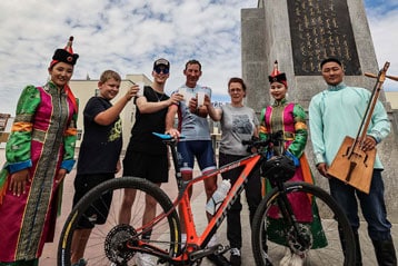Gruppe posiert mit Fahrrad vor einer Statue unter bewölktem Himmel.