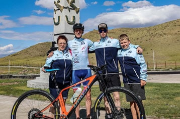 Vier Personen posieren mit einem Fahrrad vor einem hohen Denkmal unter blauem Himmel