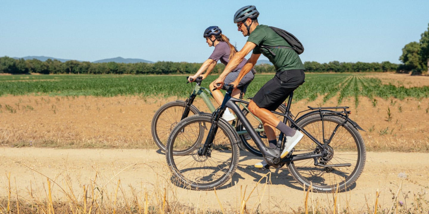 Two people cycling on a dirt path through a rural landscape.