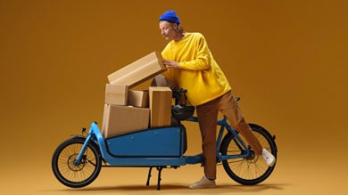 Person loading cardboard boxes onto a blue cargo bike against a mustard yellow background