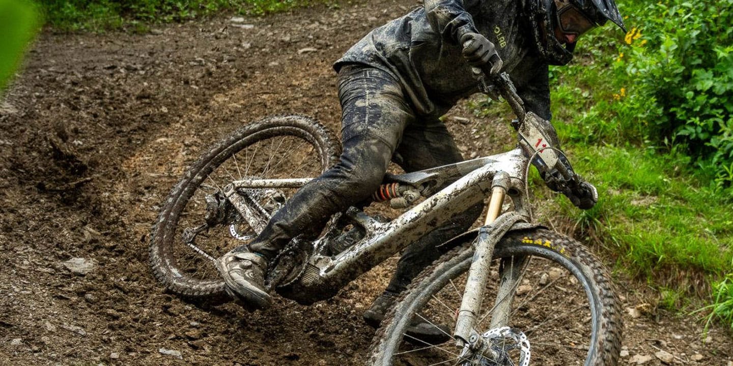 A cyclist covered in mud riding aggressively through a muddy trail in the forest.