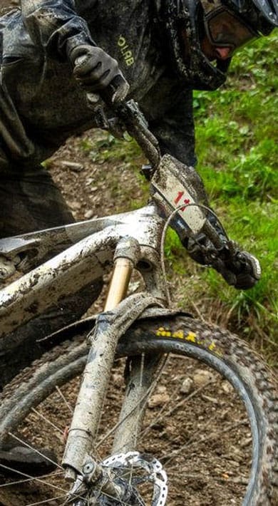 A cyclist covered in mud riding aggressively through a muddy trail in the forest.