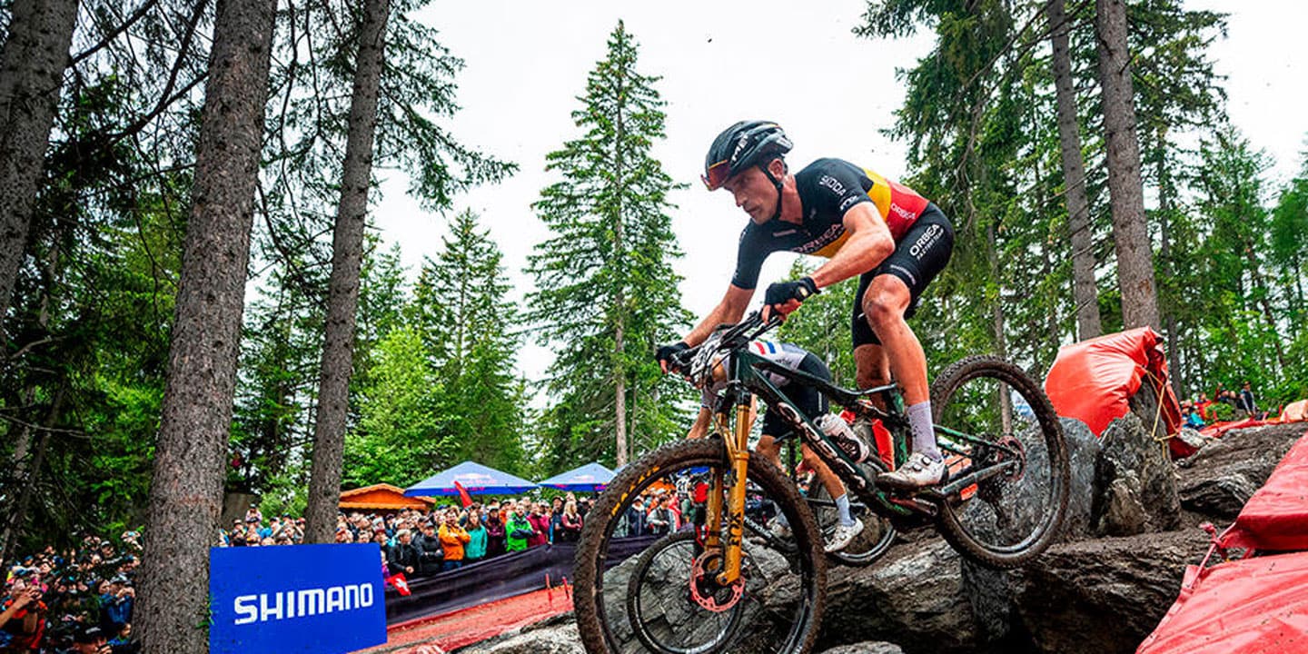 Cyclists navigating a rocky trail during a mountain bike race in a forest with spectators watching.