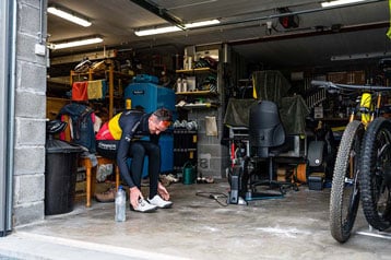 Fietser trekt schoenen aan in een garage met fietsen en gereedschap.
