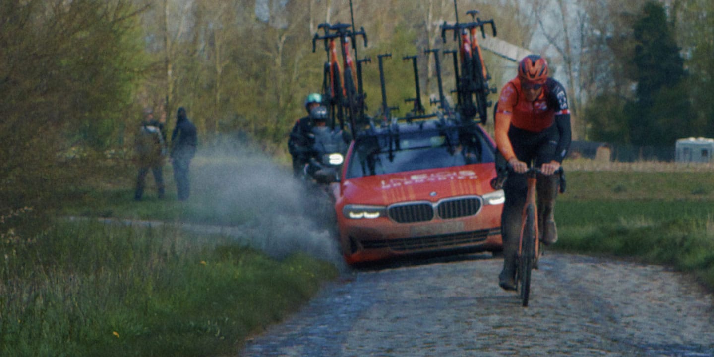 Cyclist riding on a cobblestone road followed by a support car with several bikes attached.