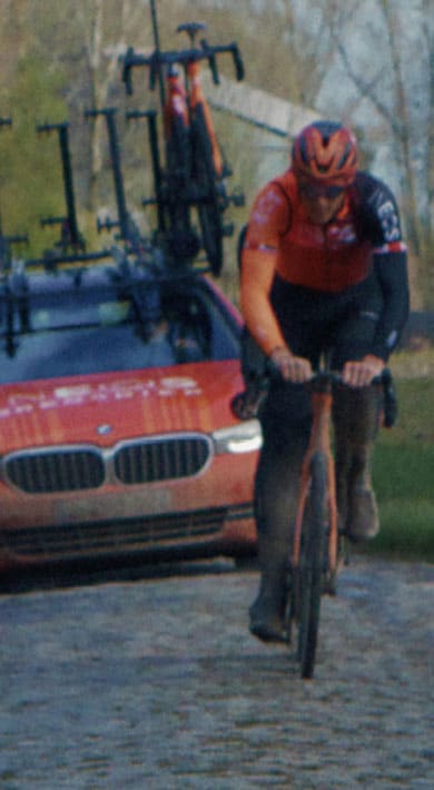 Cyclist riding on a cobblestone road followed by a support car with several bikes attached.