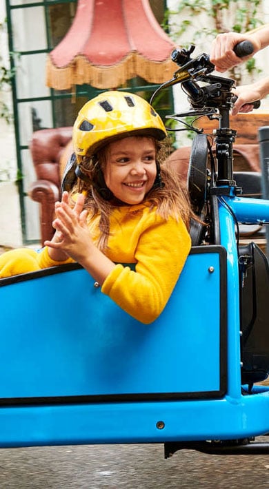 Child in a blue cargo bike, wearing a yellow outfit and helmet, smiling and clapping hands