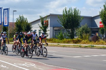 Gruppe von Radfahrern fährt an einem sonnigen Tag auf einer Straße neben einem Gebäude mit Bäumen.