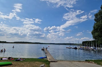 Menschen genießen einen See mit einem Dock und Segelbooten unter einem blauen Himmel.