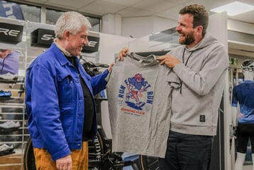 Zwei Männer lächeln und halten ein 'Run Forrest Run'-T-Shirt in einem Sportgeschäft.
