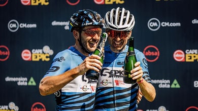 Zwei Radfahrer feiern mit Champagnerflaschen, in Radkleidung und mit Helmen