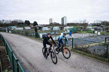 Twee fietsers rijden op een pad naast volkstuintjes.