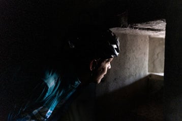 Persoon met helm kijkt door een klein raam in een schemerige ruimte.