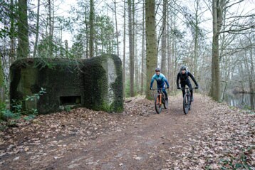 Twee fietsers rijden op een bospad bij een met mos bedekte structuur.